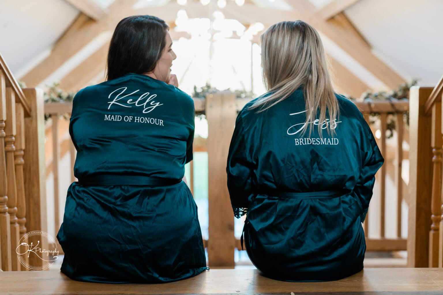 Mythe Barn Wedding photography Two women wearing robes with "Maid of Honour" and "Bridesmaid" embroidered on the back sit on wooden stairs at Mythe Barn wedding venue.