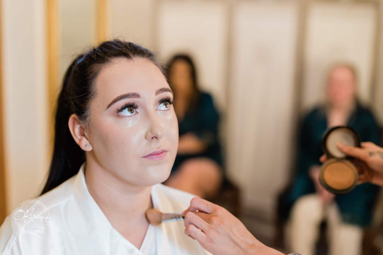 Mythe Barn Wedding photography A bride having makeup applied to her face, looking slightly upwards, with blurred bridesmaids in the background.