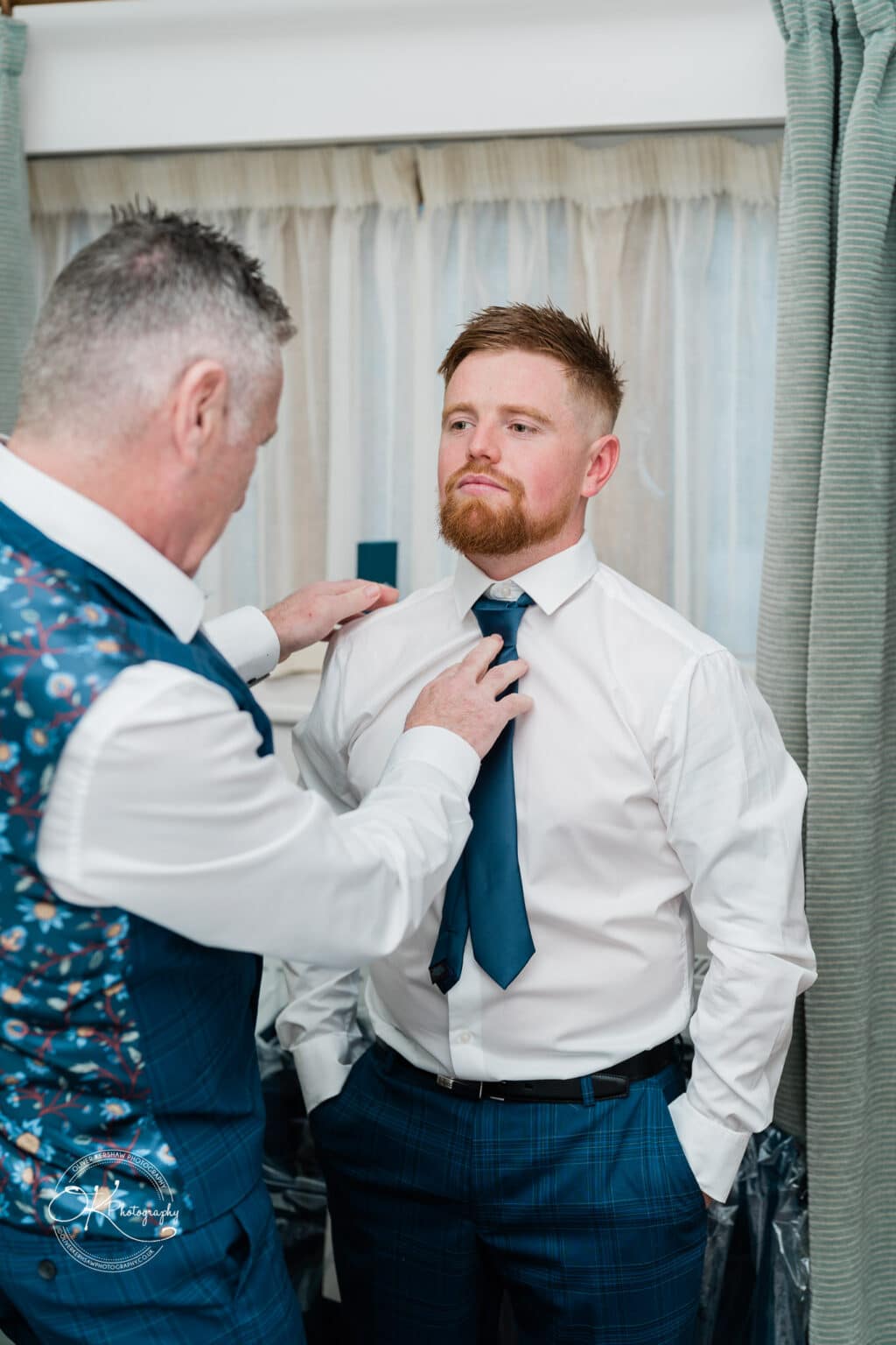 Mythe Barn Wedding photography A man adjusts the tie of another man, who is wearing a white shirt and blue checked trousers, in a room with green curtains.