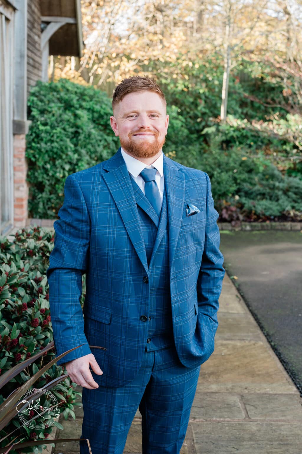 Mythe Barn Wedding photography A man in a blue checked suit and tie standing outdoors with greenery and a brick building in the background.