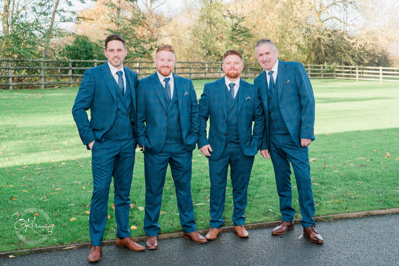 Mythe Barn Wedding photography Four men in matching blue tartan suits and brown shoes standing on a path in a grassy area with a wooden fence and trees in the background.