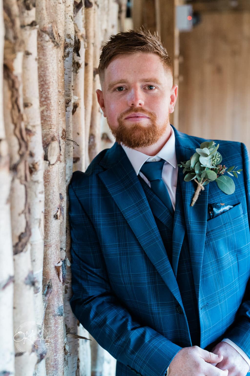 Mythe Barn Wedding photography Man in a blue plaid suit with a boutonnière, leaning against a wooden wall.