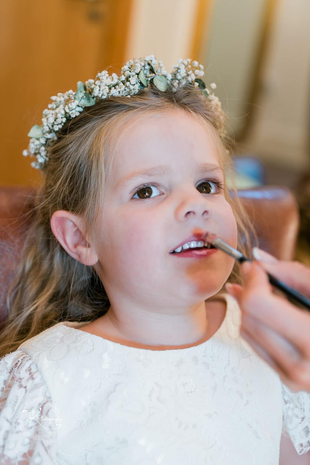 Mythe Barn Wedding photography A young girl with a flower crown has her makeup applied, wearing a white lace dress.