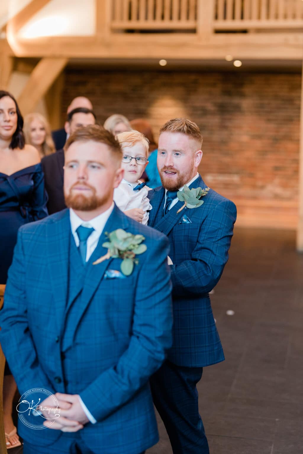 Mythe Barn Wedding photography Man in a blue checked suit with floral boutonnière stands in front of another man holding a child at a wedding ceremony in a rustic barn setting.