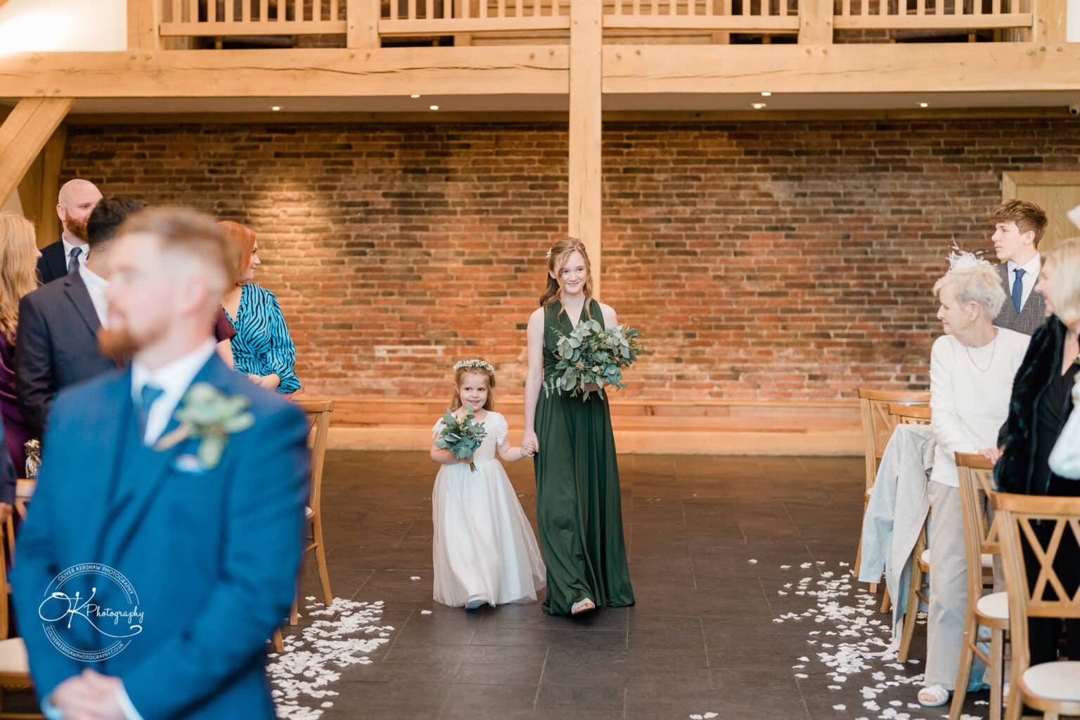 Mythe Barn Wedding photography Two flower girls walk down the aisle at a wedding in Mythe Barn, one in a white dress and the other in a dark green dress, holding bouquets of greenery.