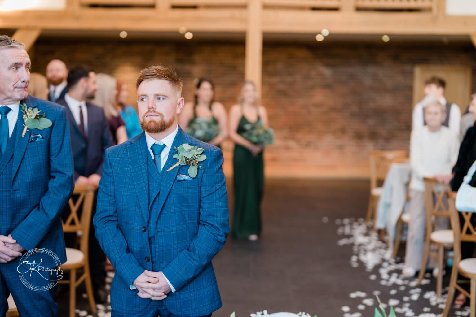 Mythe Barn Wedding photography Two men in blue suits with boutonnières stand at the front of a wedding venue, with guests seated behind them.