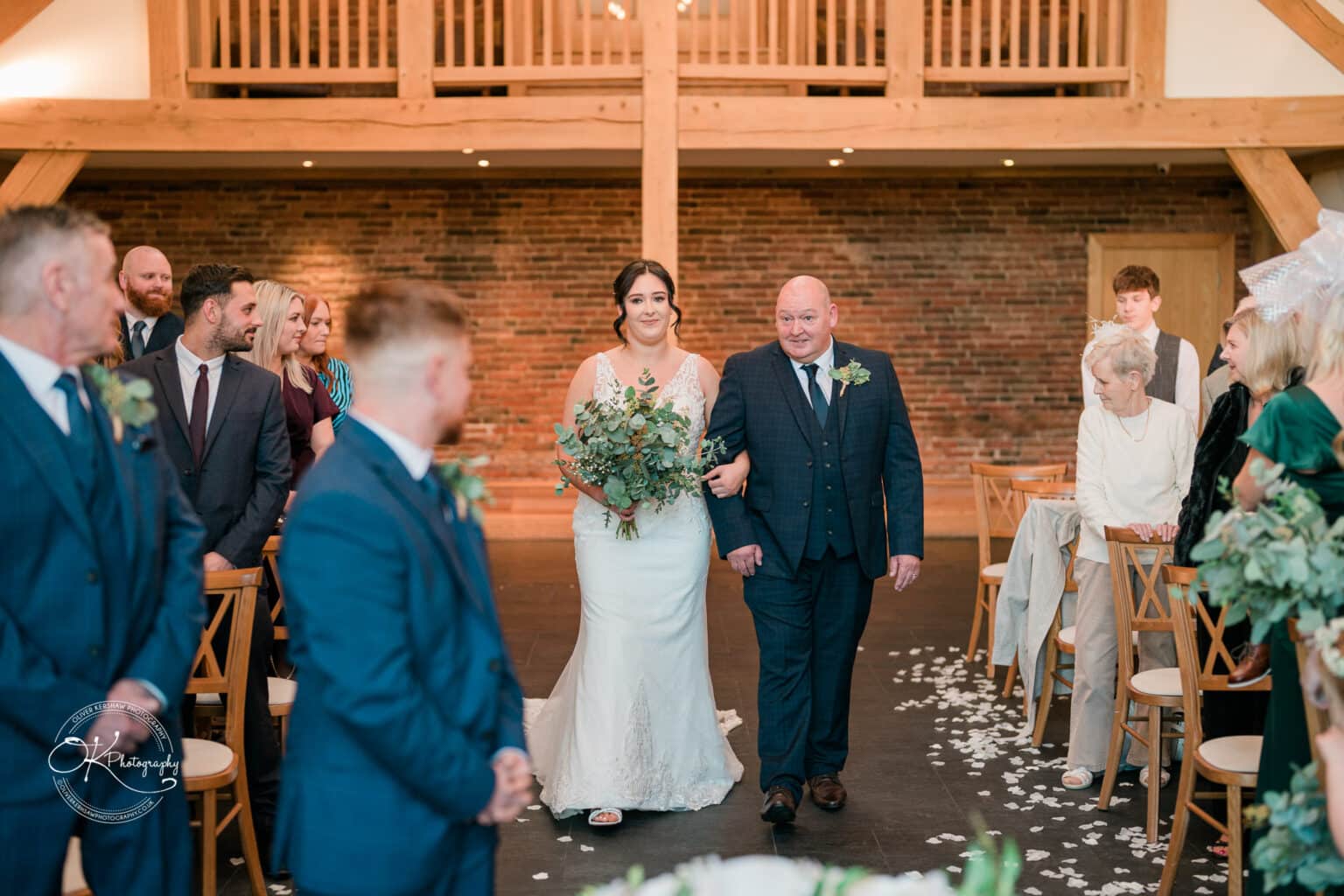 Mythe Barn Wedding photography A bride walks down the aisle with her father in a rustic barn wedding venue, with guests seated on either side watching the procession.