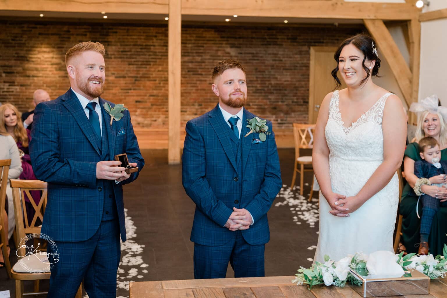 Mythe Barn Wedding photography A bride in a white dress stands with two groomsmen in blue plaid suits holding a ring box, smiling during a wedding ceremony at Mythe Barn.