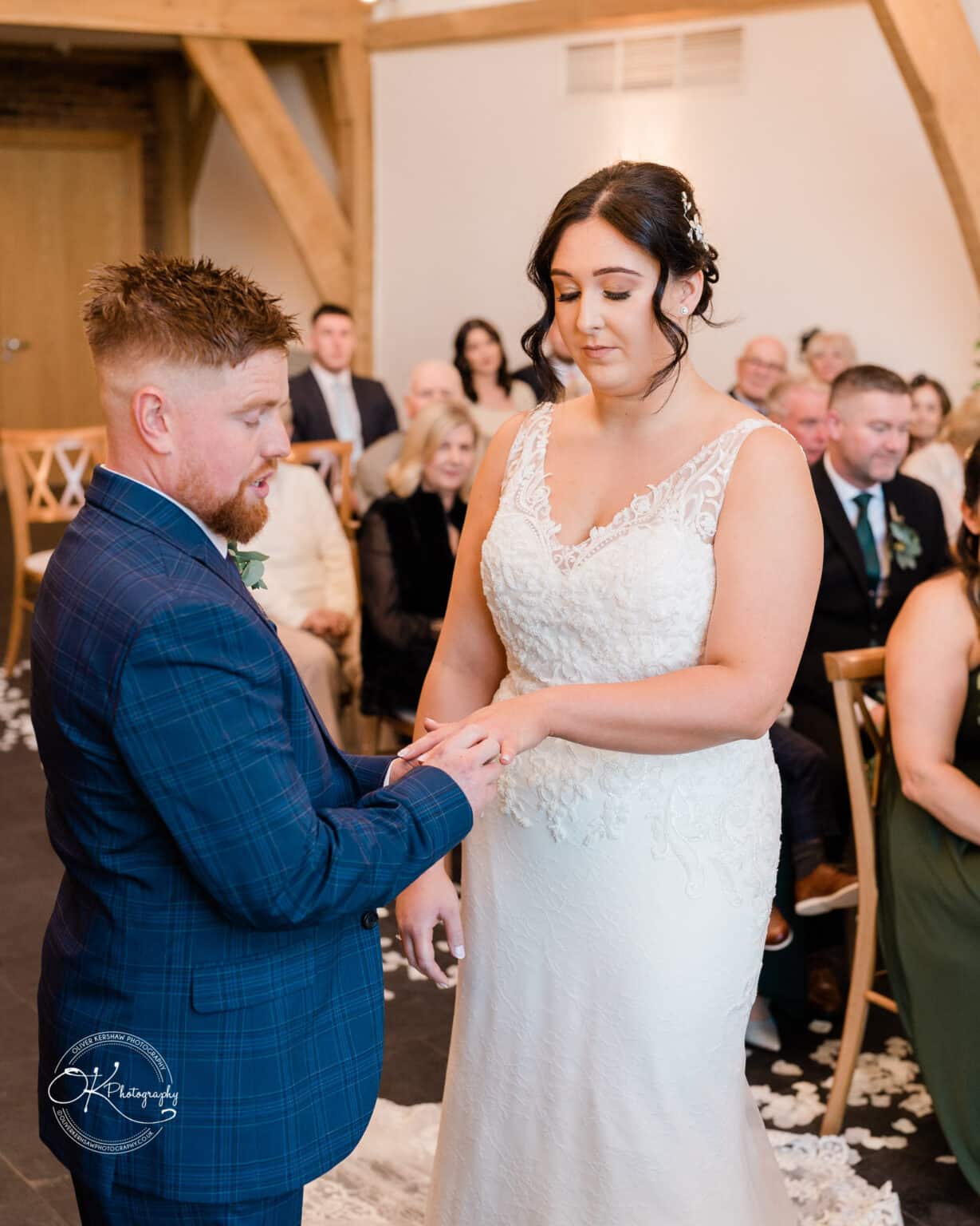 Mythe Barn Wedding photography A groom in a blue suit placing a ring on the bride's finger in a wooden-beamed venue during their wedding ceremony with guests seated in the background.
