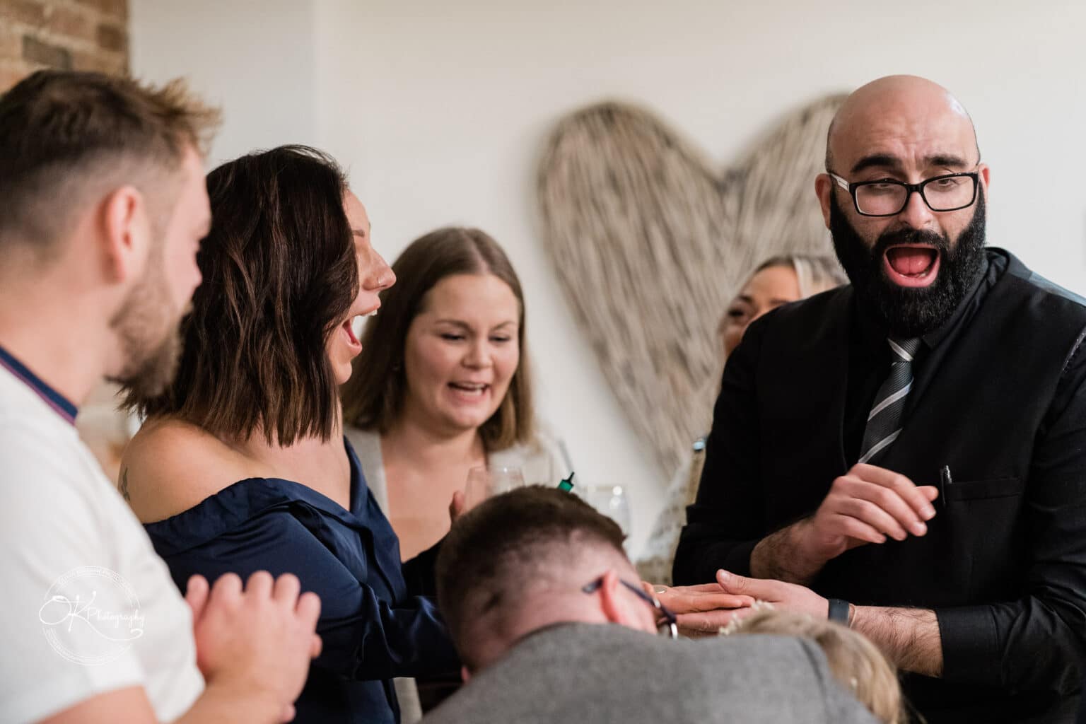 Mythe Barn Wedding photography-By-Oliver-Kershaw-Photography-OK902958 A group of people laughing and reacting to a man in a black suit entertaining them at a wedding.