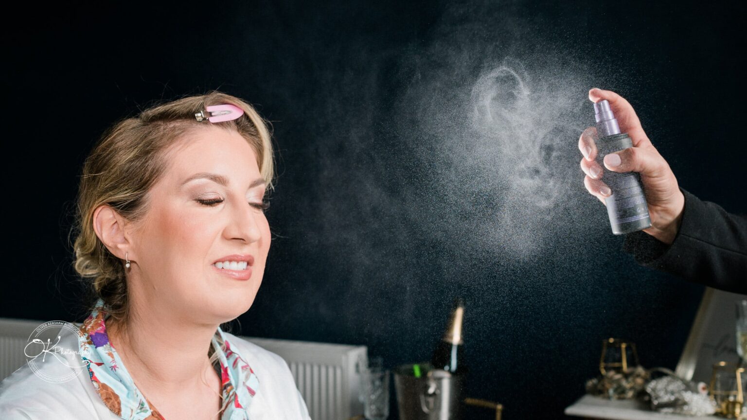 A woman with eyes closed, having her hair done with a pink clip, while a hand sprays a mist in front of her face.
