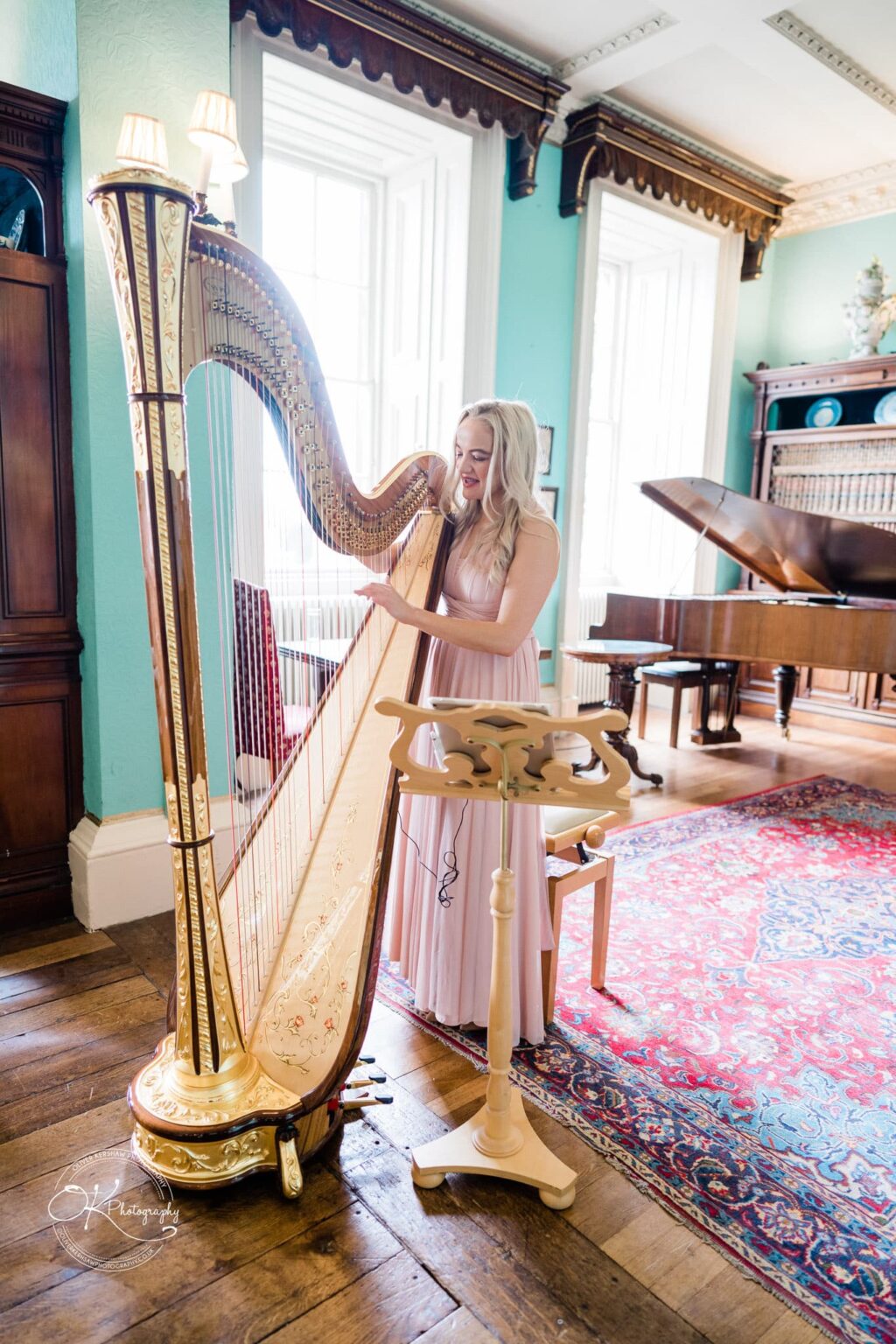 A woman playing a golden harp in an elegant room with large windows, a piano, wooden floors, and teal walls at Prestwold Hall.