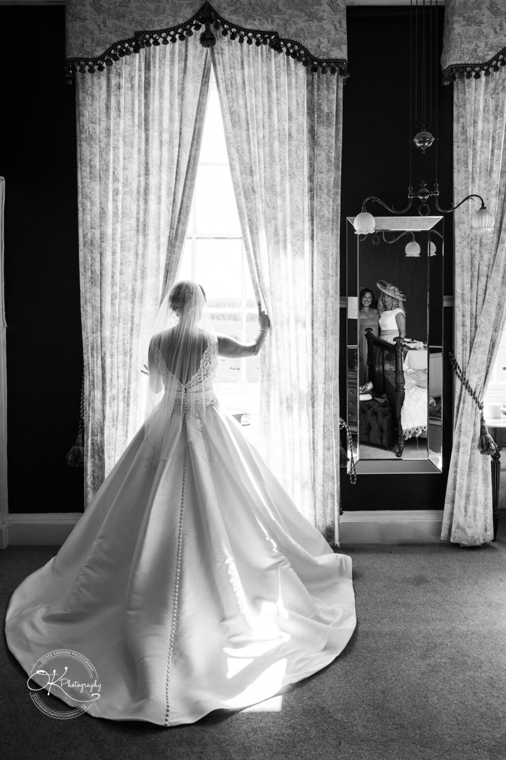 Bride standing by a window in a room with ornate curtains, her reflection seen in a mirror behind showing her and two other women.