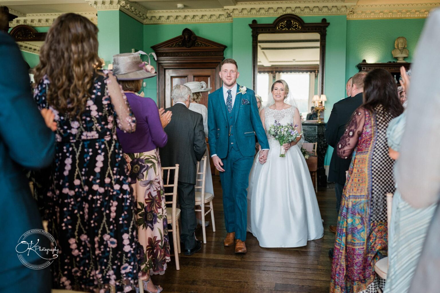 A newlywed couple walking down the aisle while guests applaud inside a grand, elegantly decorated hall with mint green walls and ornate woodwork.