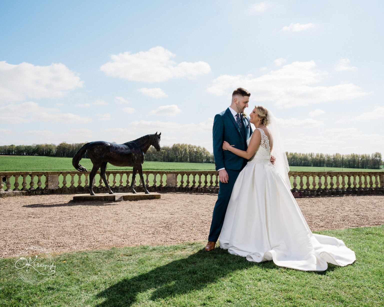 Bride and groom standing near a horse statue in the garden of Prestwold Hall, with a scenic view of fields and trees in the background.