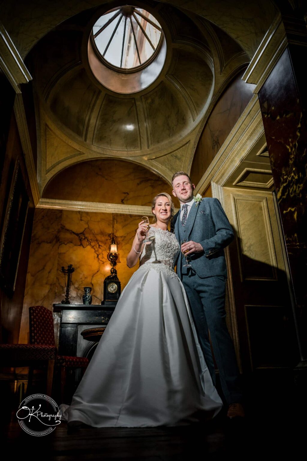 A bride and groom smiling while holding champagne glasses inside Prestwold Hall, standing under a domed ceiling.