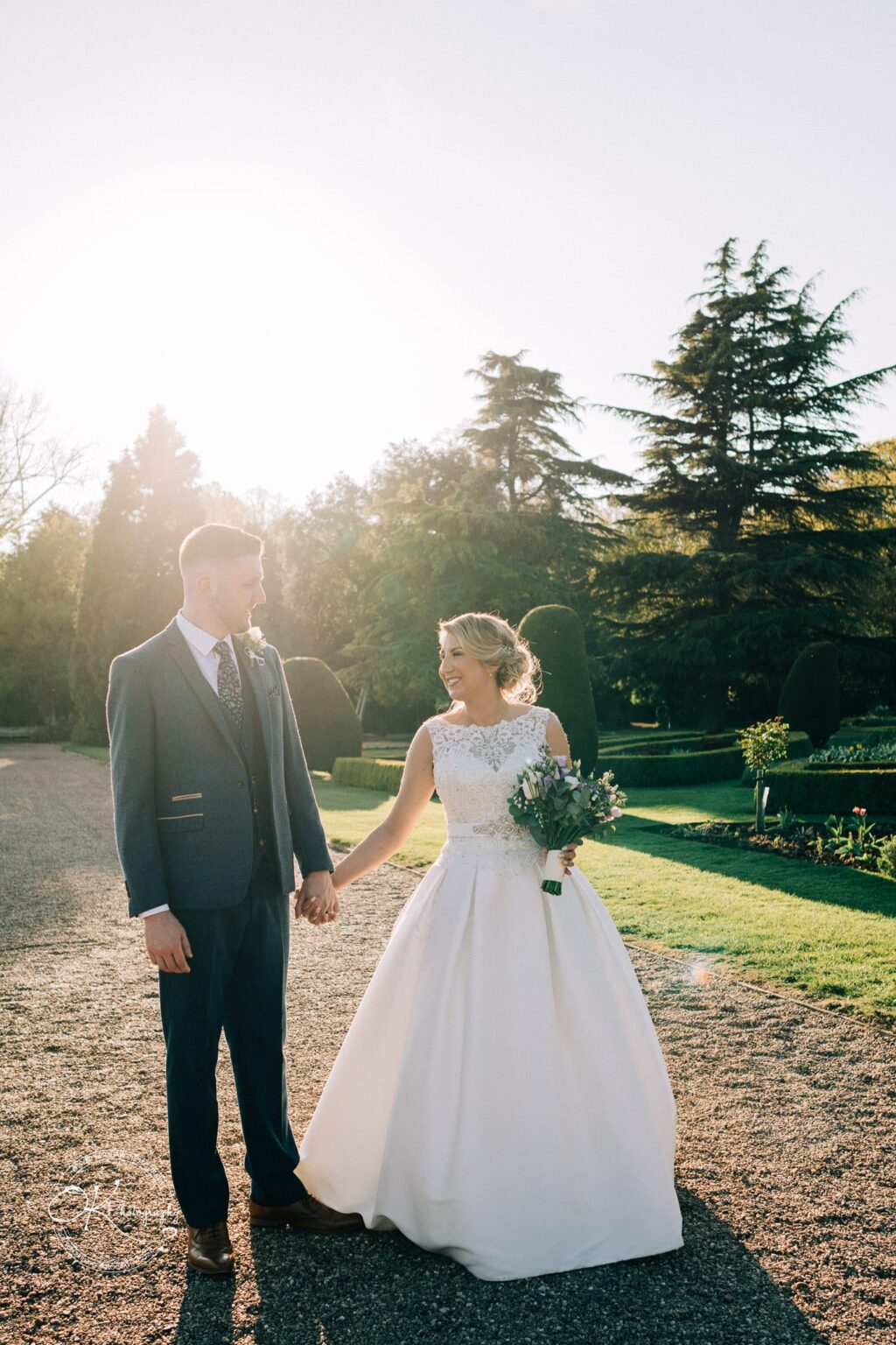A bride and groom holding hands outdoors at Prestwold Hall, with manicured gardens and trees in the background.