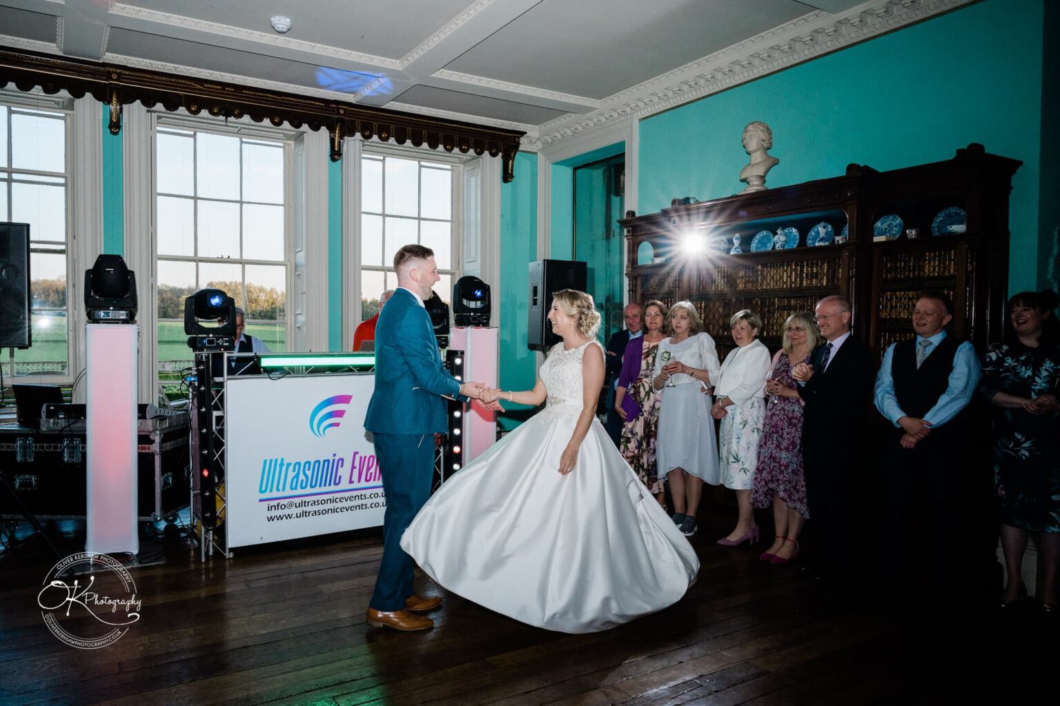A bride and groom dance in a brightly lit hall with a DJ booth in the background, surrounded by smiling guests.