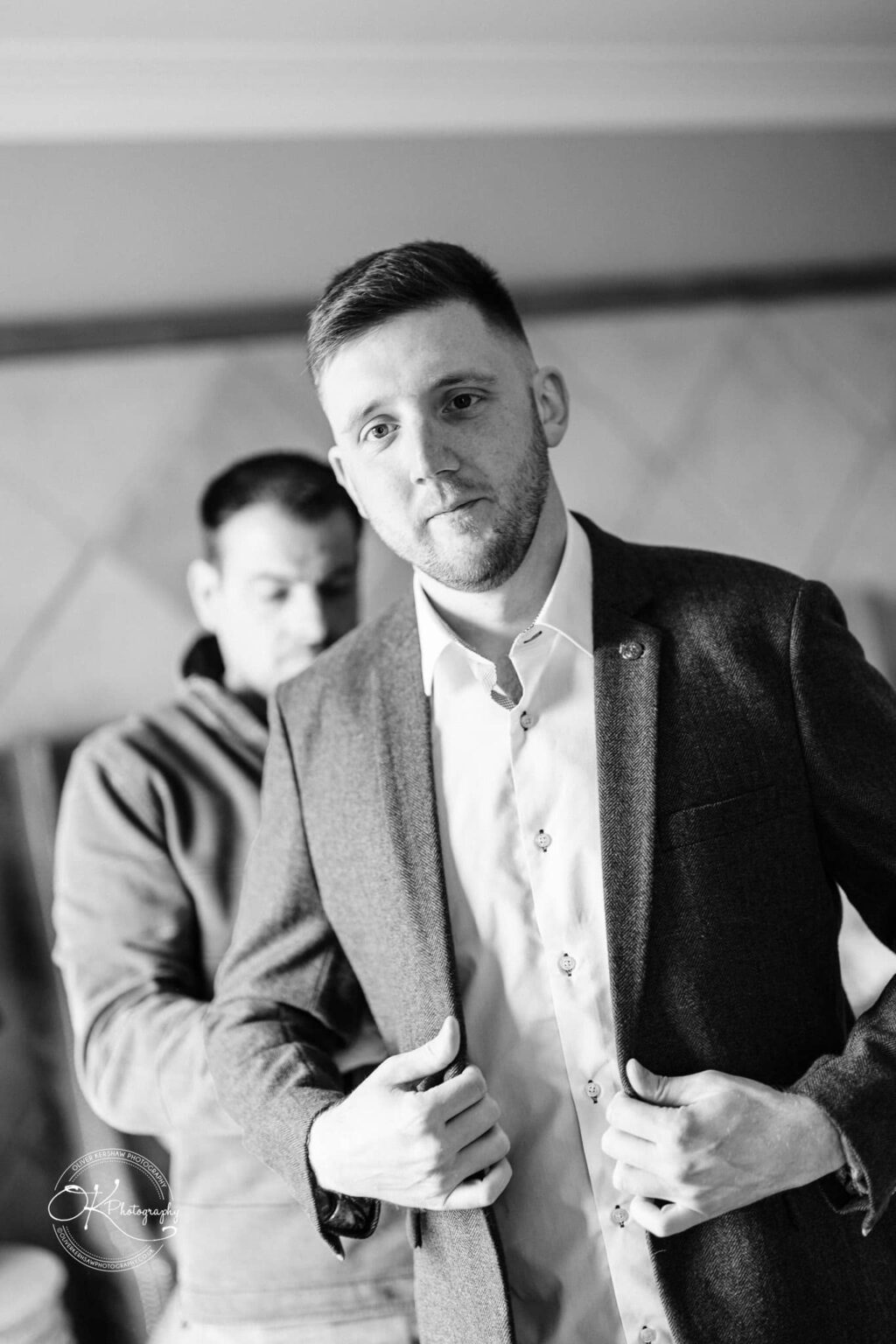 A man buttoning his suit jacket while another man stands in the background. The image is in black and white.