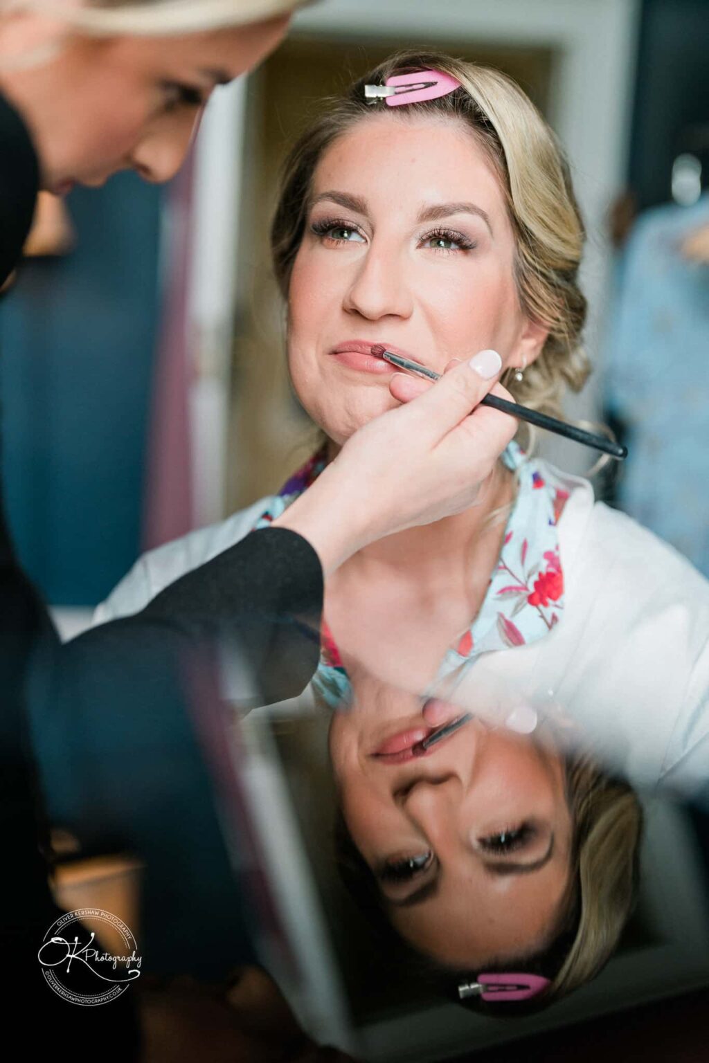 A woman is having her makeup applied, with her reflection visible in a mirror below.