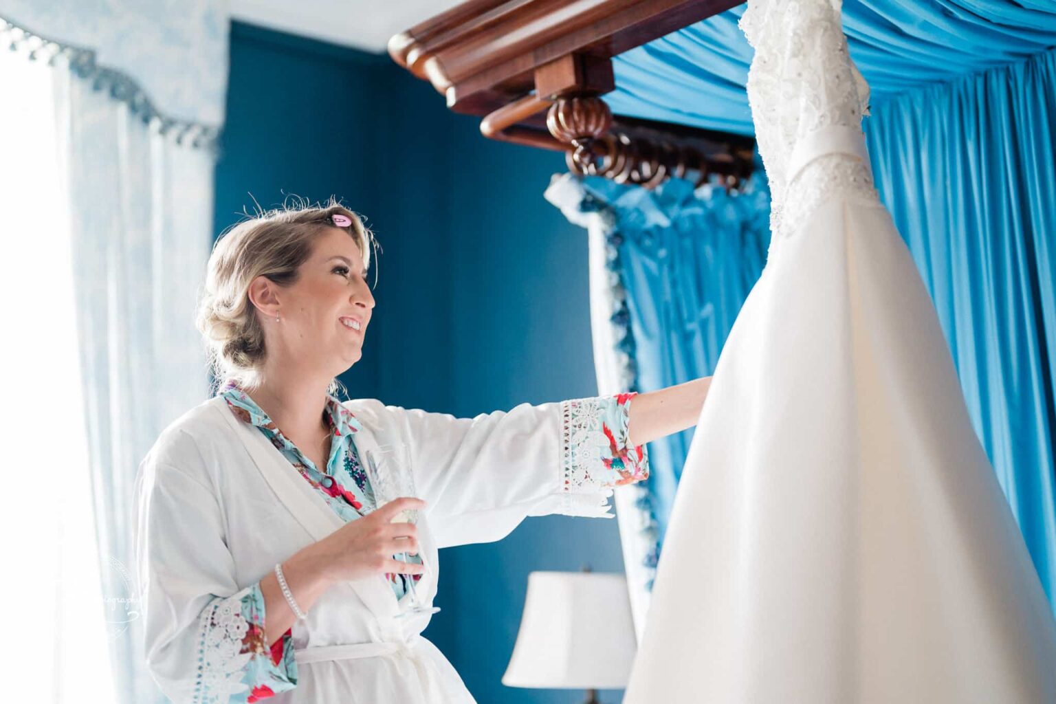 A woman in a robe smiles as she reaches for a white wedding dress hanging in a blue room with a four-poster bed.