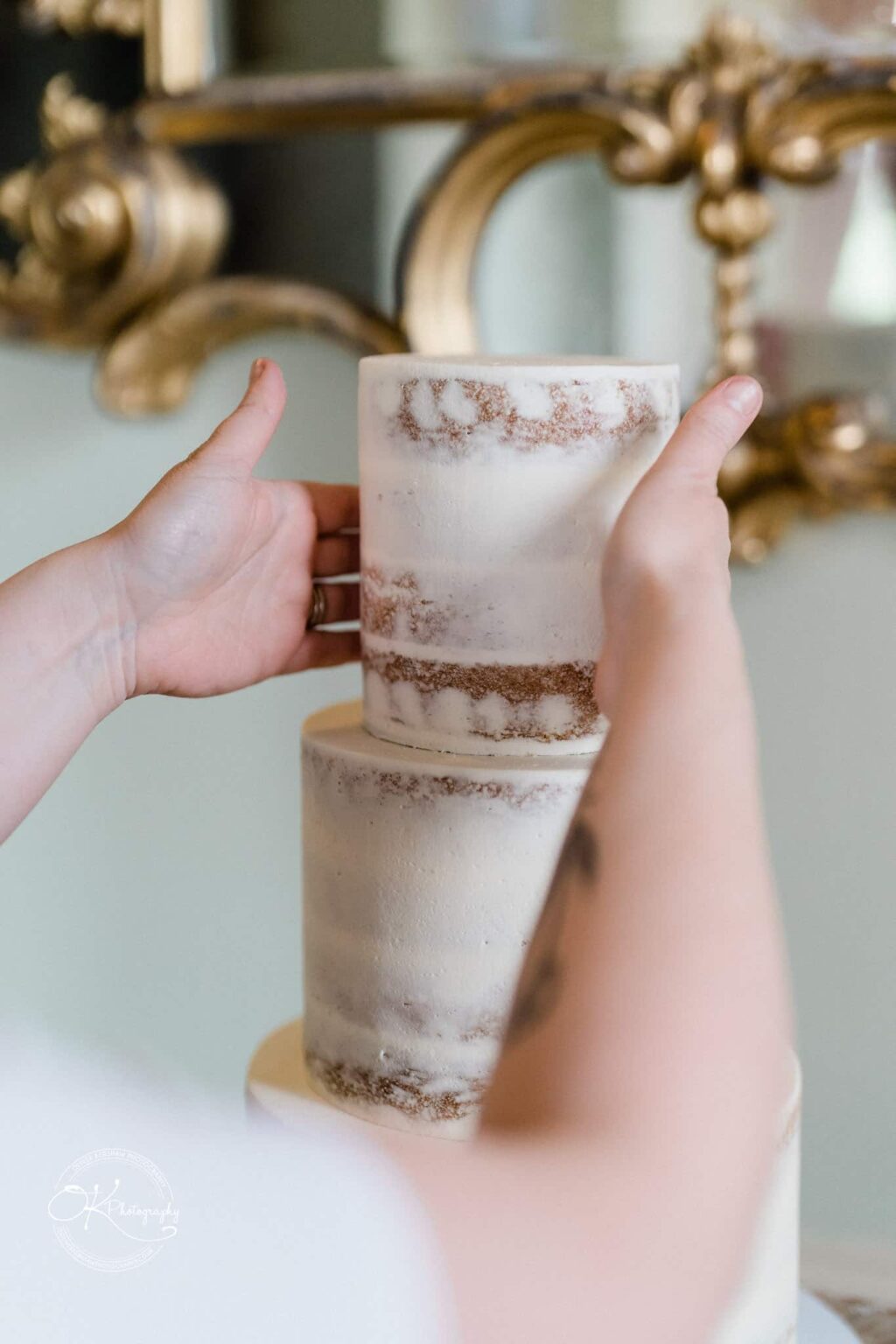 A person assembling a tiered naked cake with minimal frosting, with a gold ornate mirror in the background.