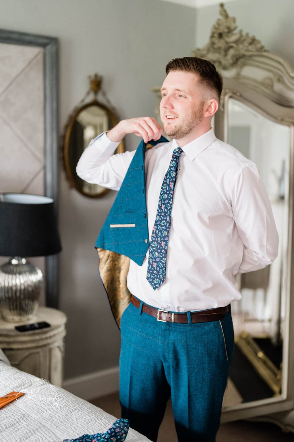 A man in formal attire putting on a blue suit in an elegantly decorated room with vintage mirrors and furniture.