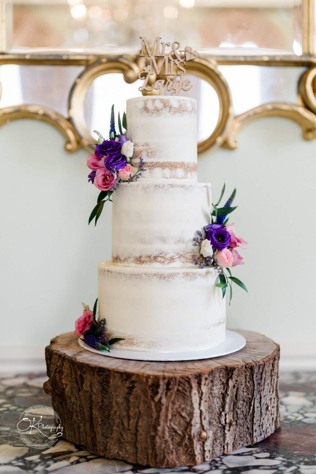 Three-tier semi-naked wedding cake decorated with purple and pink flowers, topped with a "Mr & Mrs Large" cake topper, displayed on a wooden base.