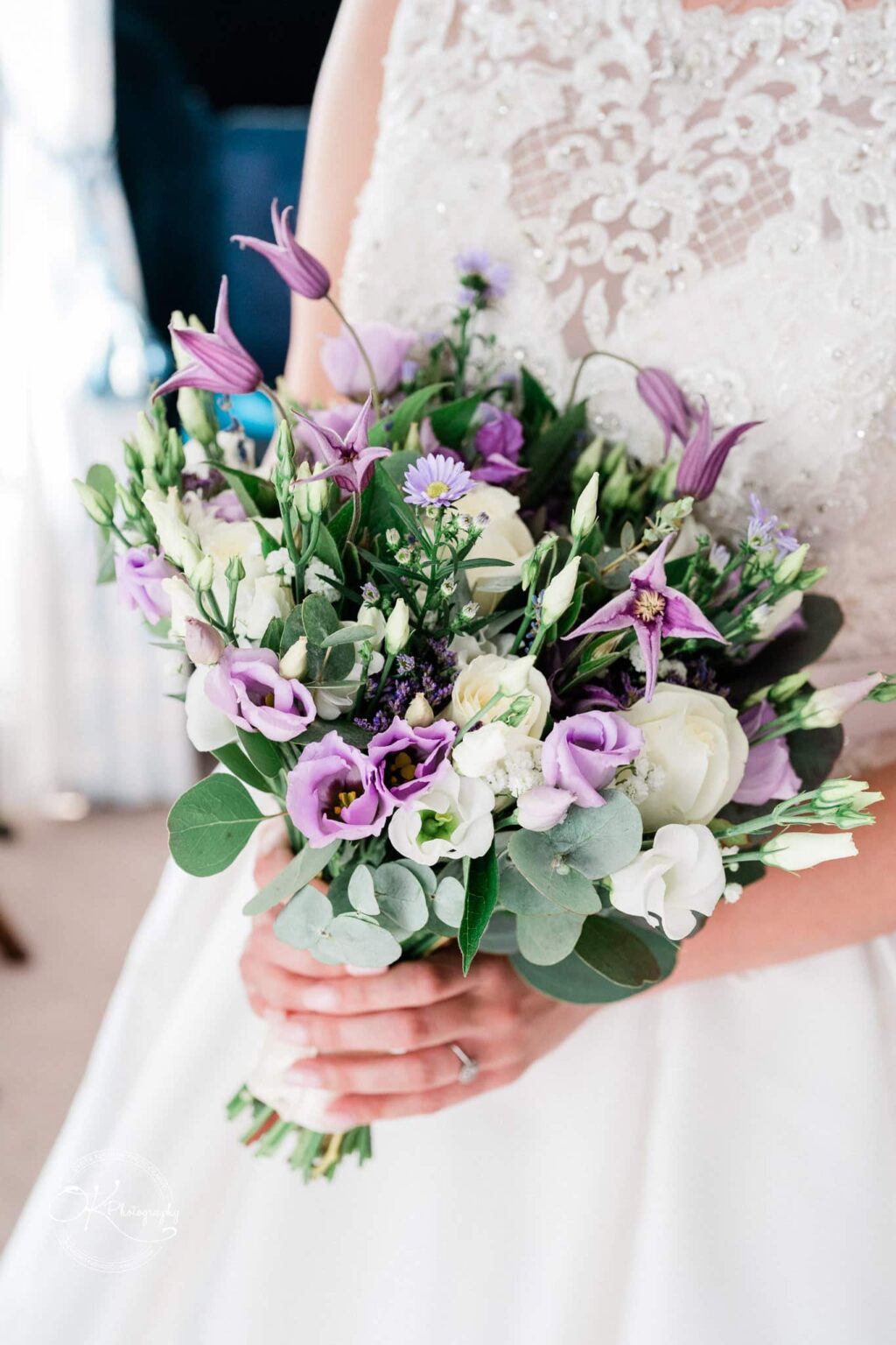 A bride holding a bouquet of purple and white flowers with greenery, wearing a white lace wedding dress.