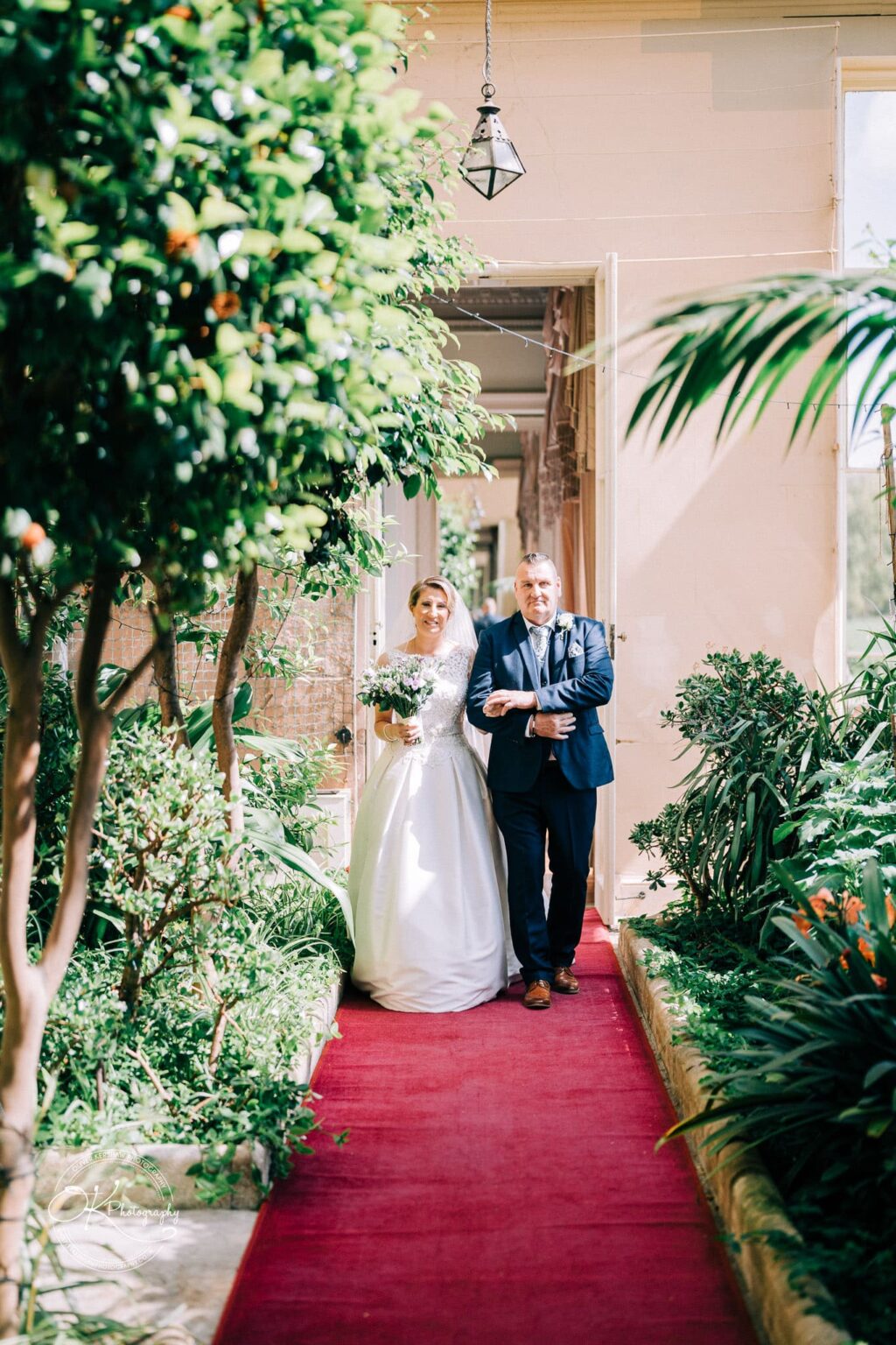 A bride and a man walk down a red carpeted aisle surrounded by greenery and trees at Prestwold Hall.