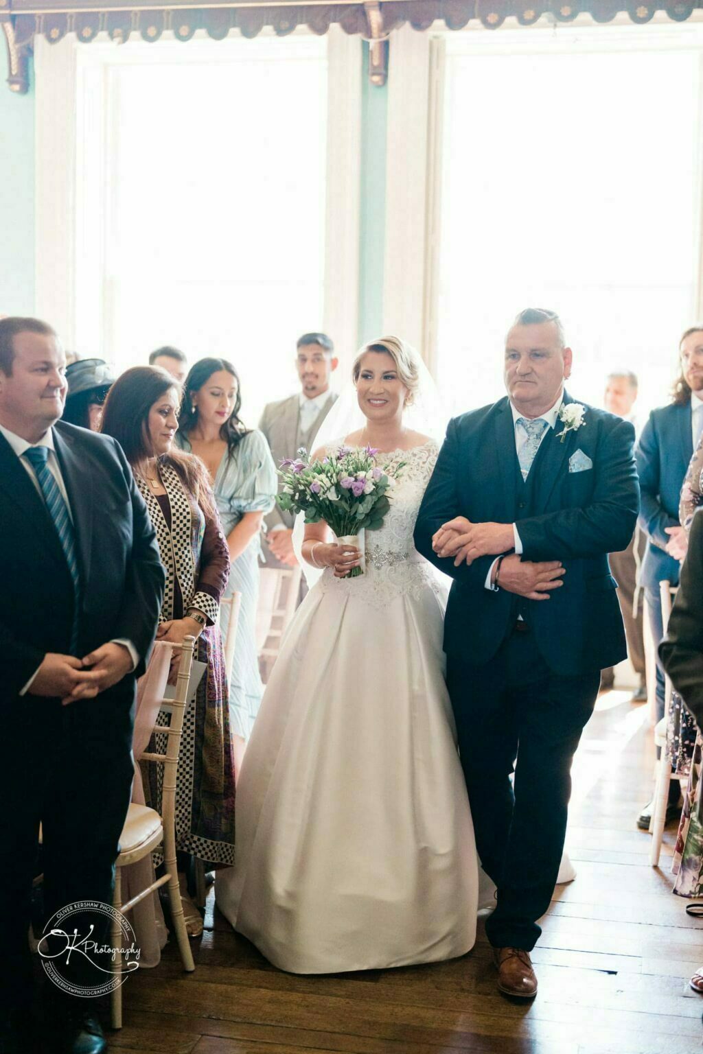 Bride in white dress holding flowers, walking down the aisle with a man in a dark suit, surrounded by wedding guests at Prestwold Hall.