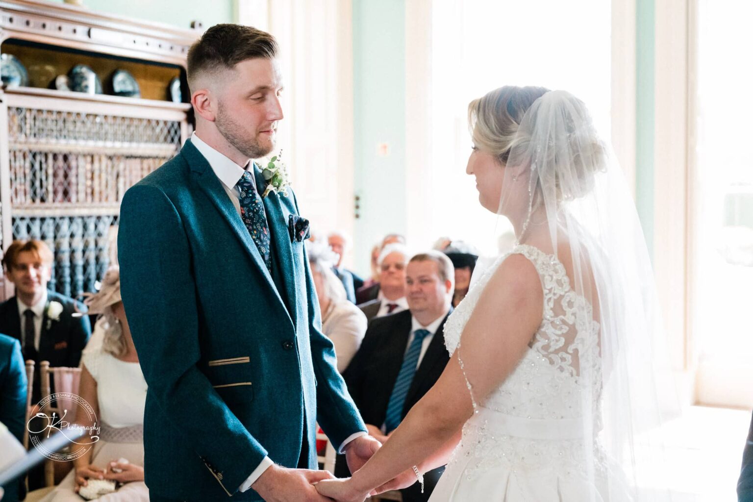 A bride and groom holding hands and facing each other during their wedding ceremony at Prestwold Hall, with seated guests in the background.