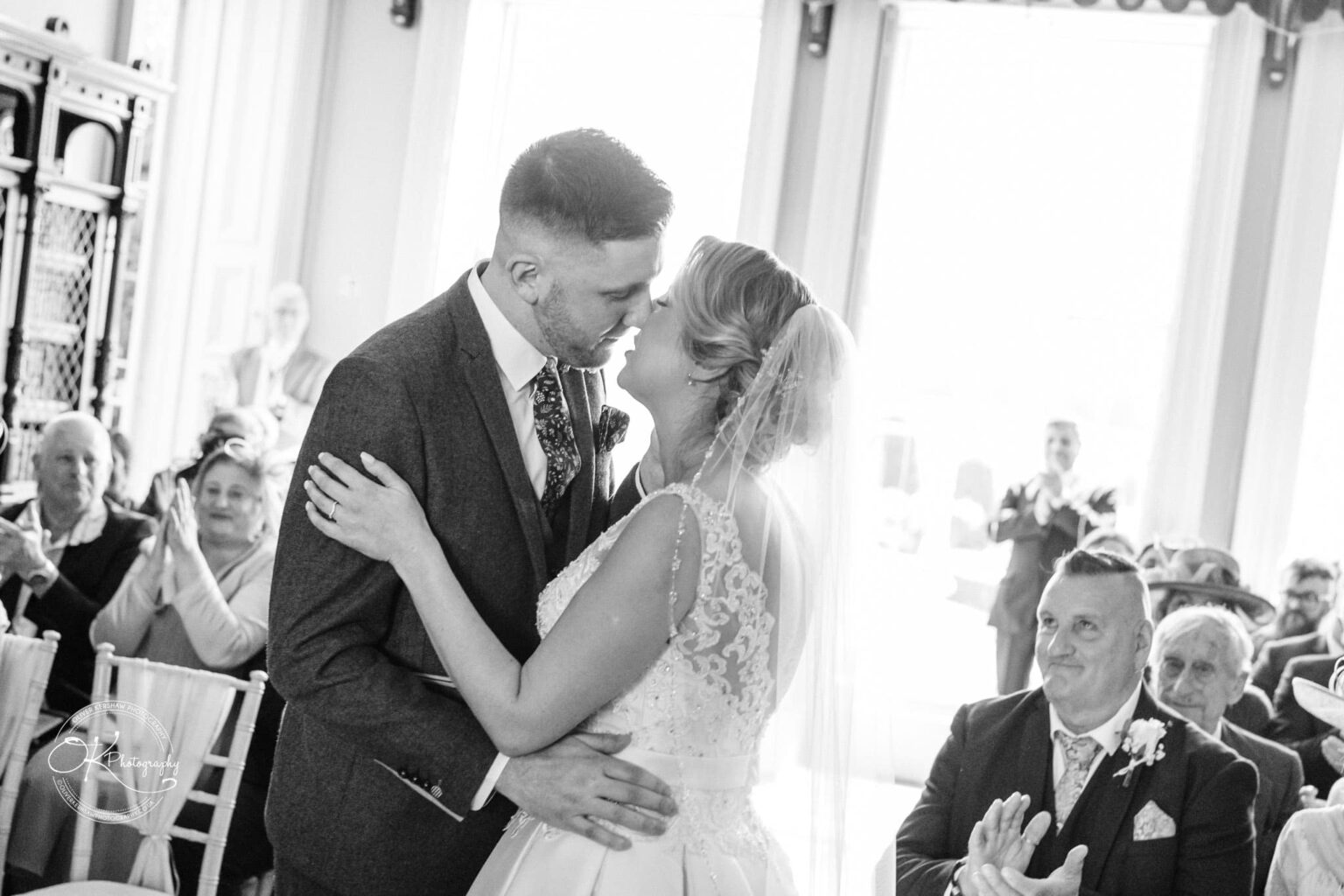 A newlywed couple kissing during their wedding ceremony while guests applaud in the background.