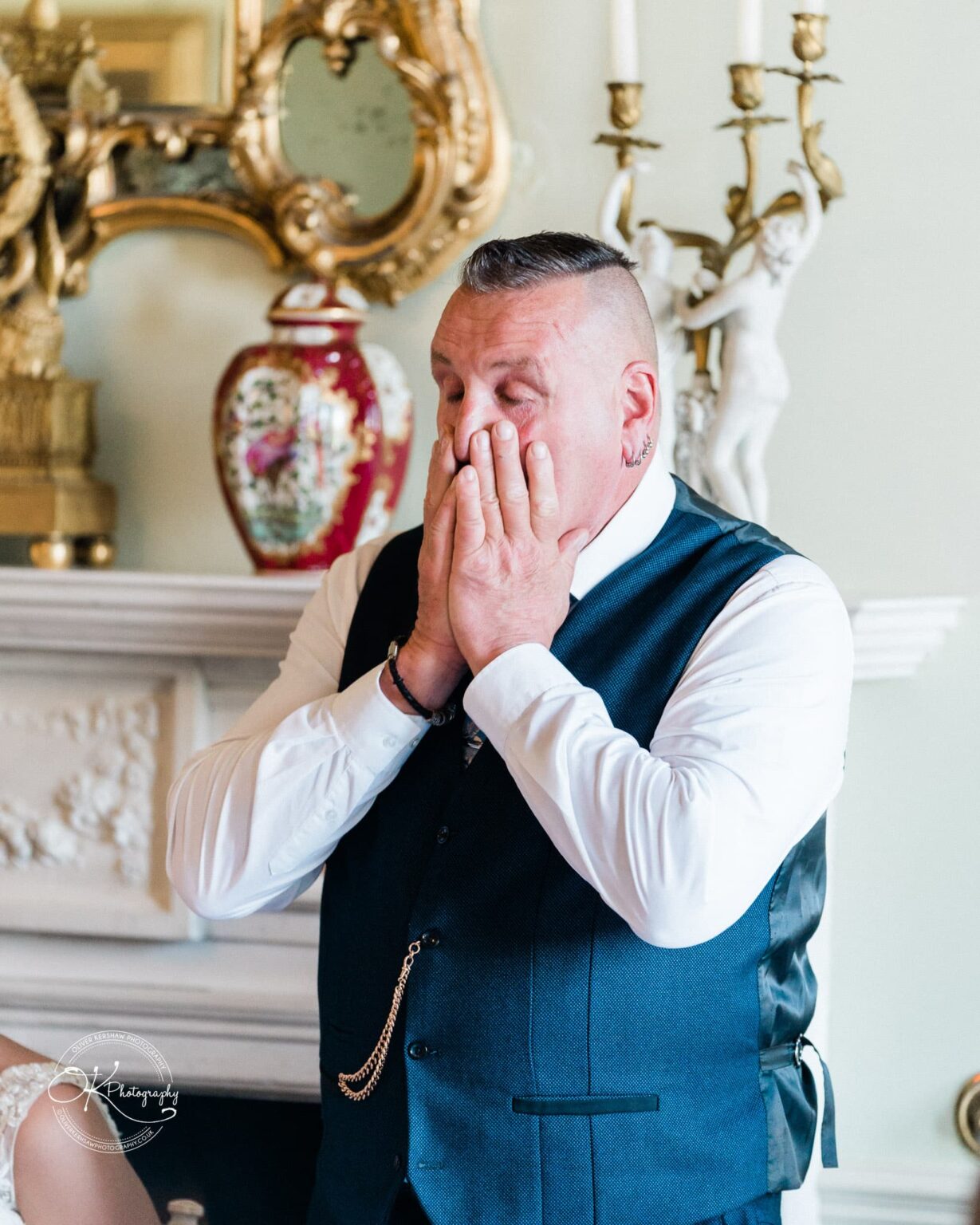 A man in a vest standing indoors with his hands covering his face in an emotional moment. Detailed decor is visible in the background including a candelabra, vase, and ornate mirror.