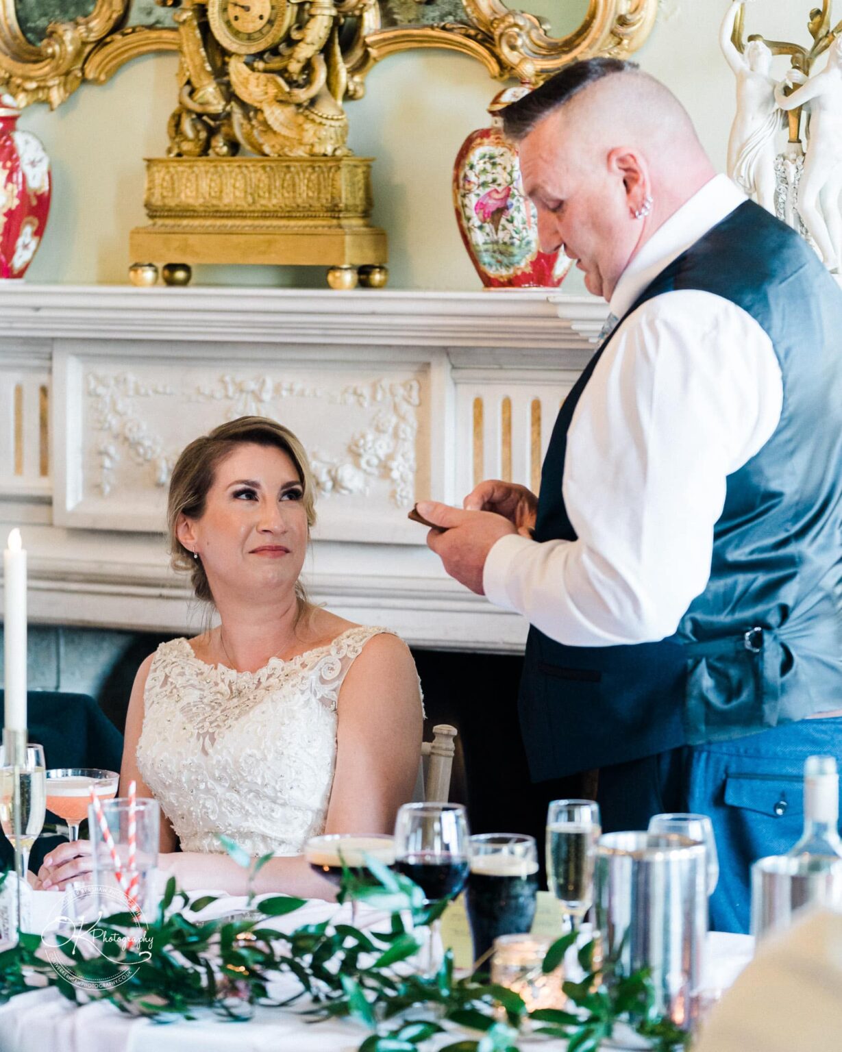 A bride in a white wedding dress sits at a table looking up at a man in a vest, who is holding a piece of paper, in an elegantly decorated room with a fireplace and ornate gold decorations.