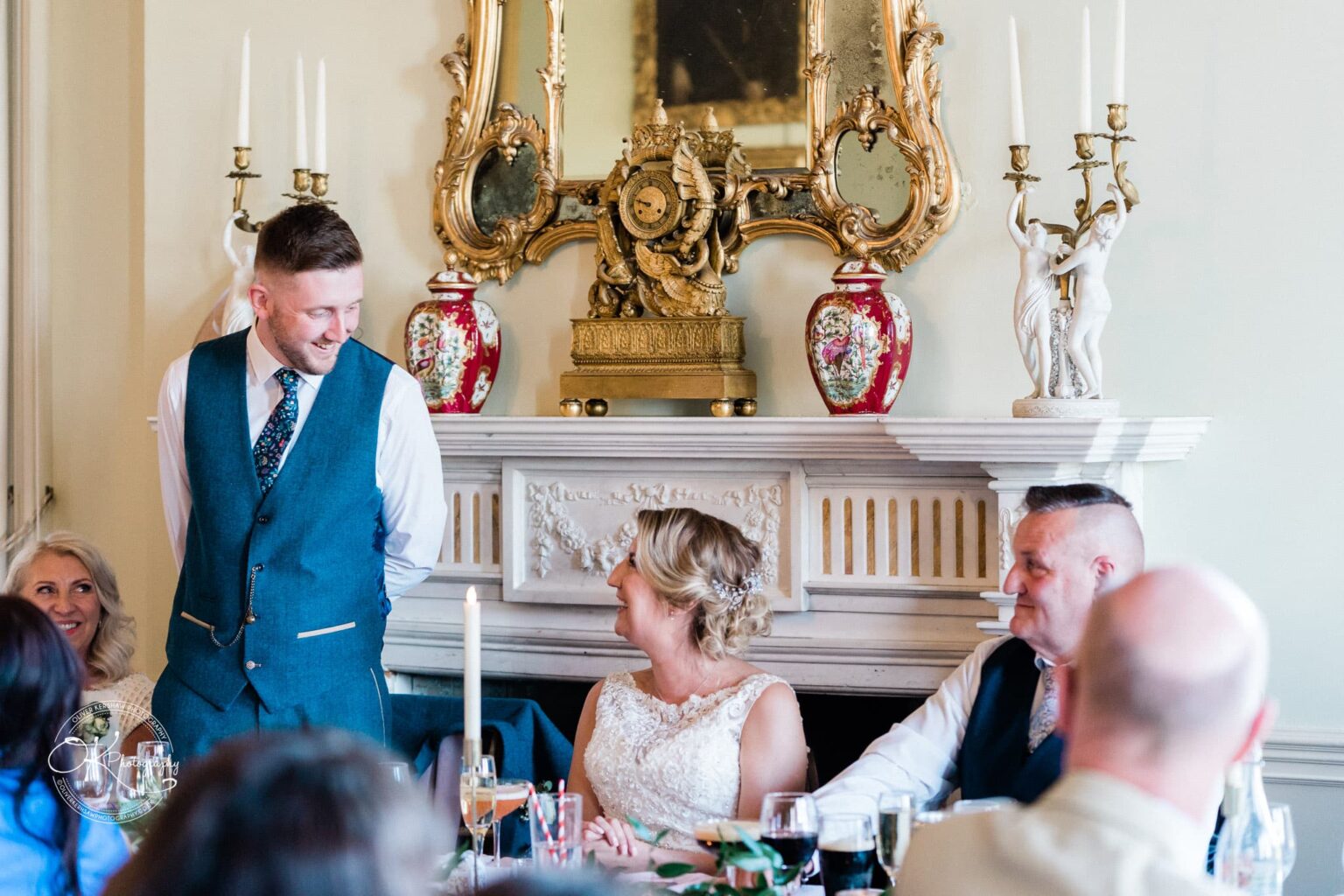 A wedding speech being given in an elegant room with an ornate fireplace decorated with red vases, candelabras, and a golden clock.