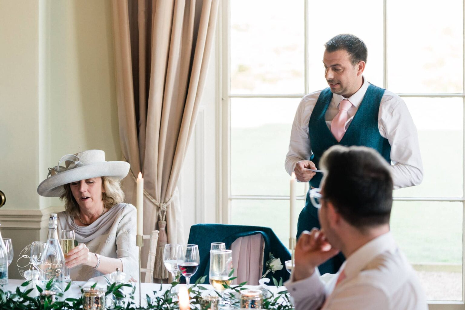 A man wearing a suit gives a speech while another woman wearing a large hat listens and holds a glass at a dining table in Prestwold Hall.