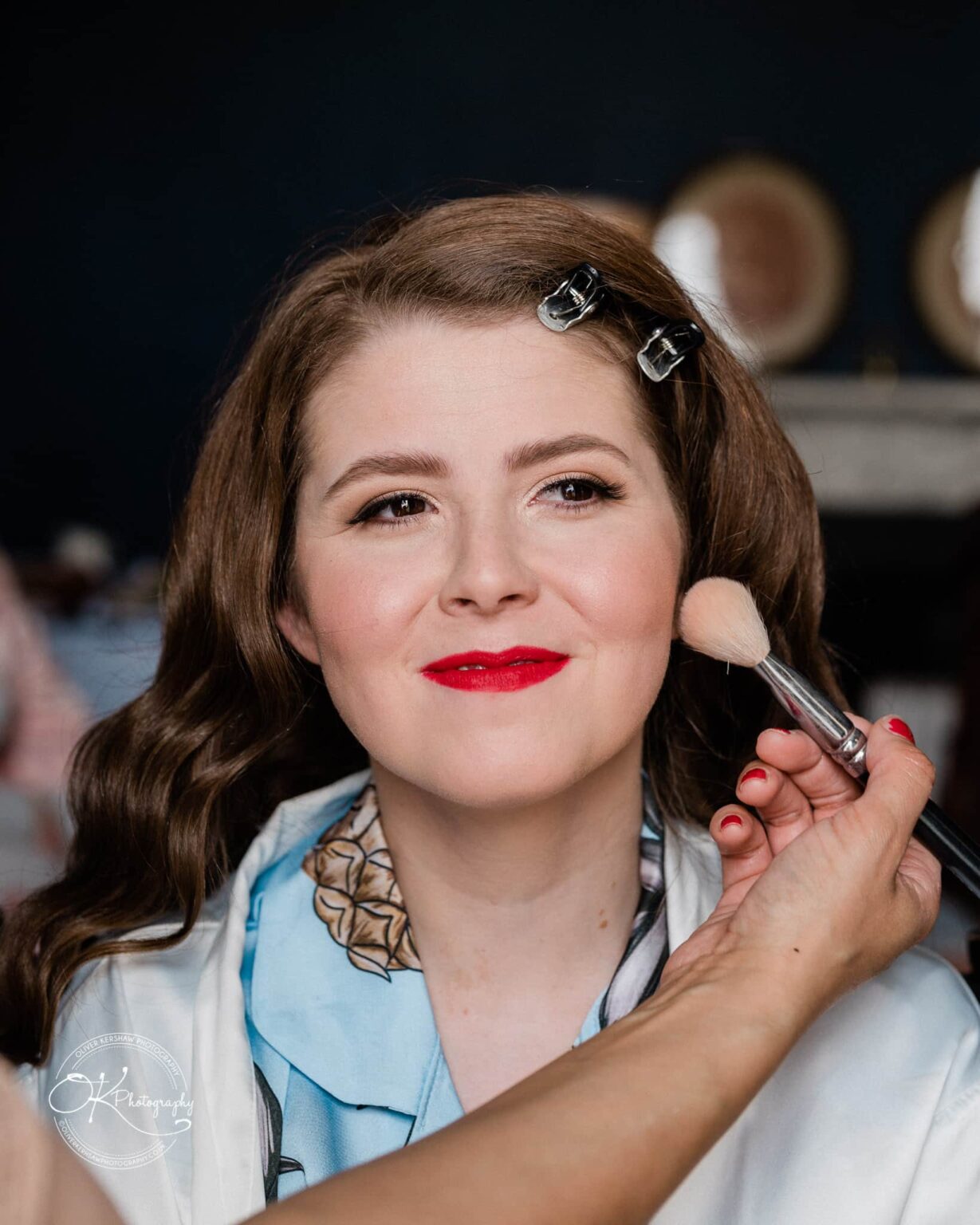 Prestwold Hall Wedding Photography A woman having makeup applied with a brush, wearing a light blue top and hair clips.