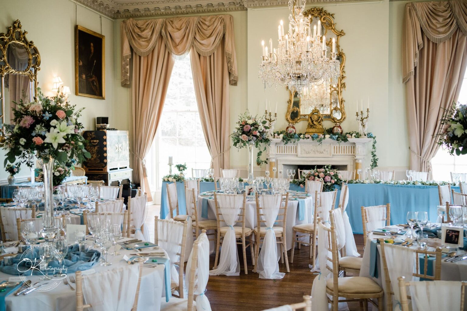 Prestwold Hall Wedding Photography Elegant dining room at Prestwold Hall set for an event, featuring round tables adorned with white and light blue tablecloths, floral centrepieces, and crystal chandeliers.