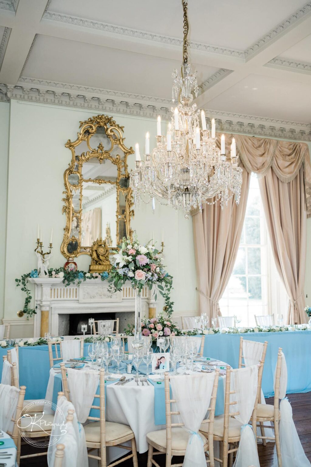 Prestwold Hall Wedding Photography Elegant dining room decorated for an event at Prestwold Hall, featuring a grand chandelier, ornate mirror, floral arrangements, and round tables set with blue tablecloths.