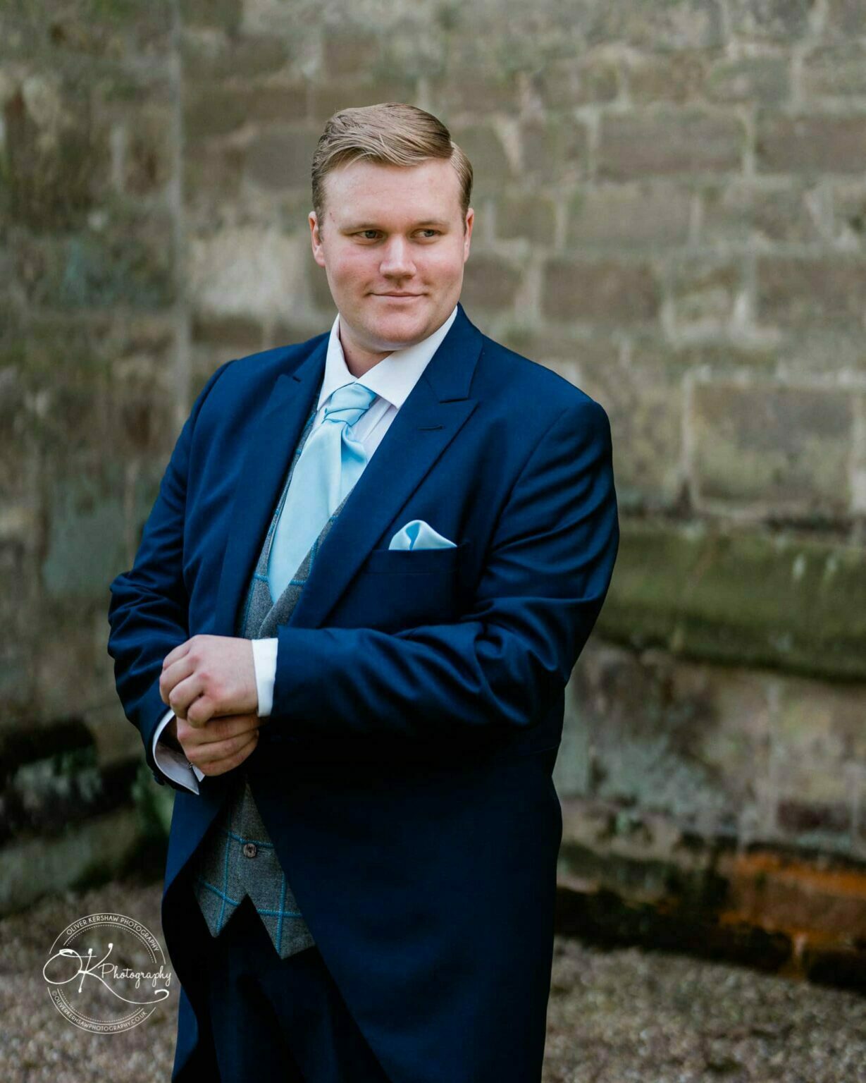 Prestwold Hall Wedding Photography A man in a blue suit stands in front of a stone wall, adjusting his cuffs, with a logo in the lower left corner that reads "OK Photography".