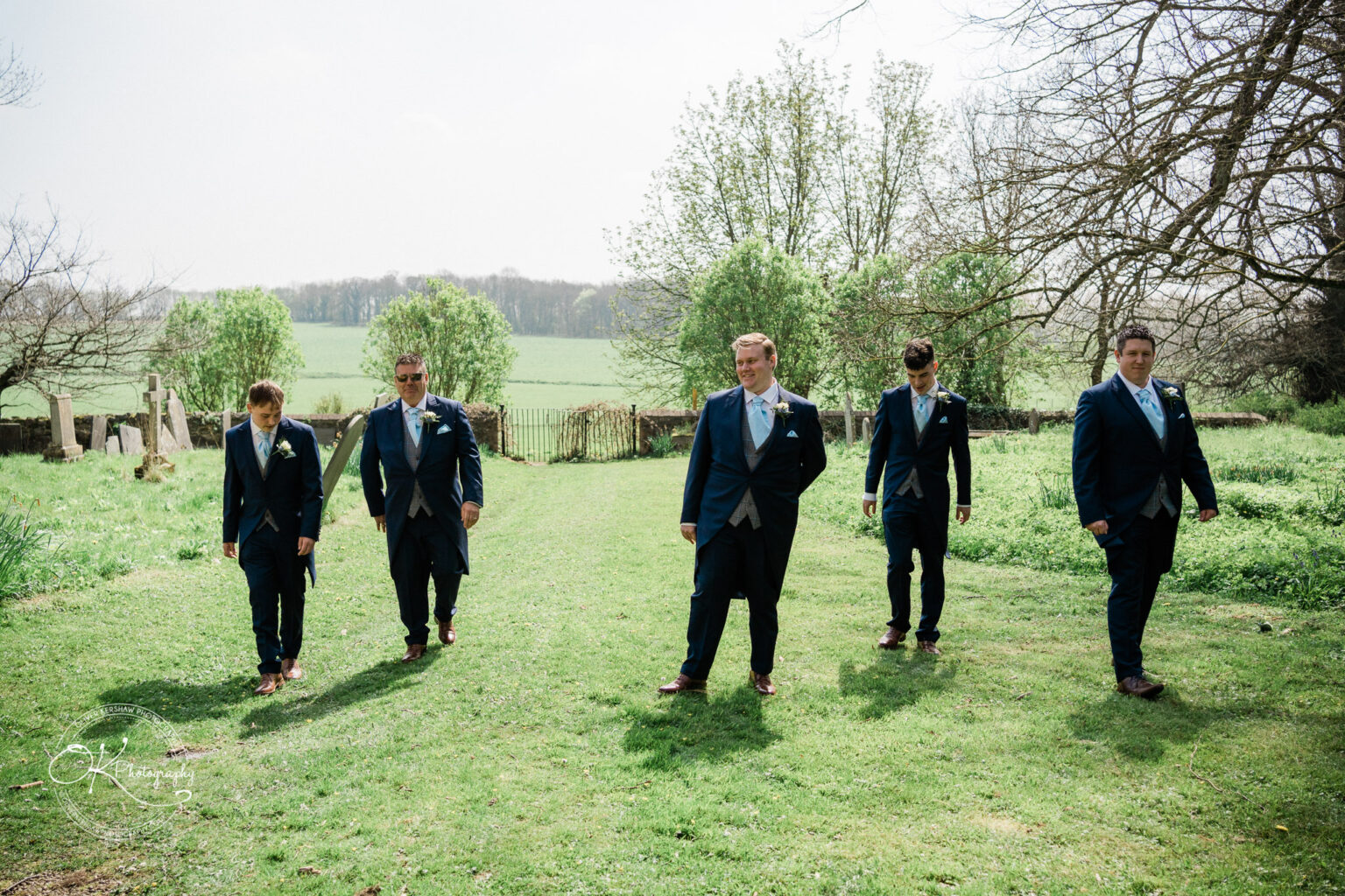 Prestwold Hall Wedding Photography Five men in formal suits walking on a grassy path, with a scenic countryside background of trees and fields.