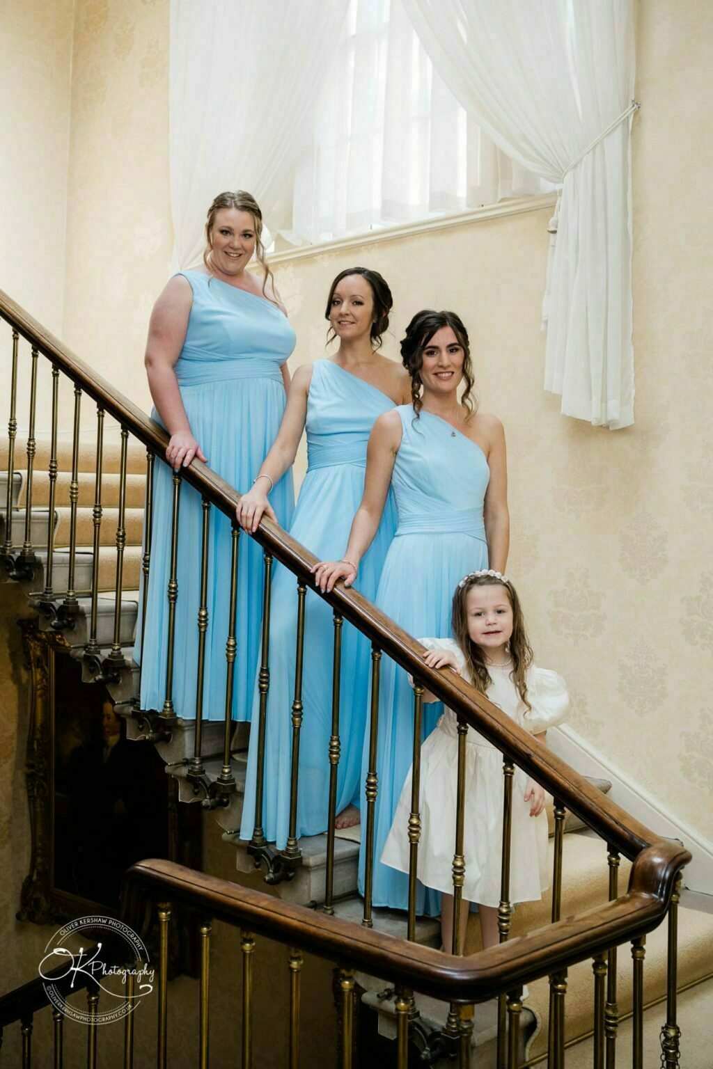 Prestwold Hall Wedding Photography Four women in light blue dresses and a young girl in a white dress standing on a staircase at Prestwold Hall.