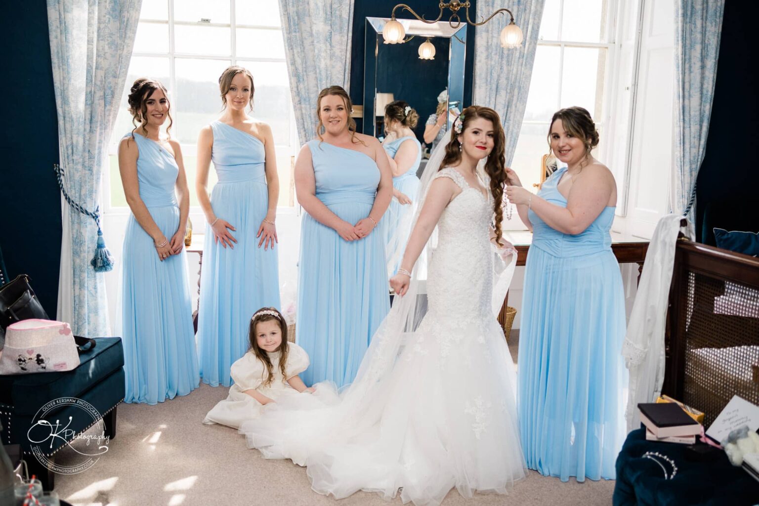 Prestwold Hall Wedding Photography A bride in a lace wedding dress, flanked by five bridesmaids in light blue dresses, poses with a young flower girl in a white dress in a well-lit room with large windows and elegant blue curtains.