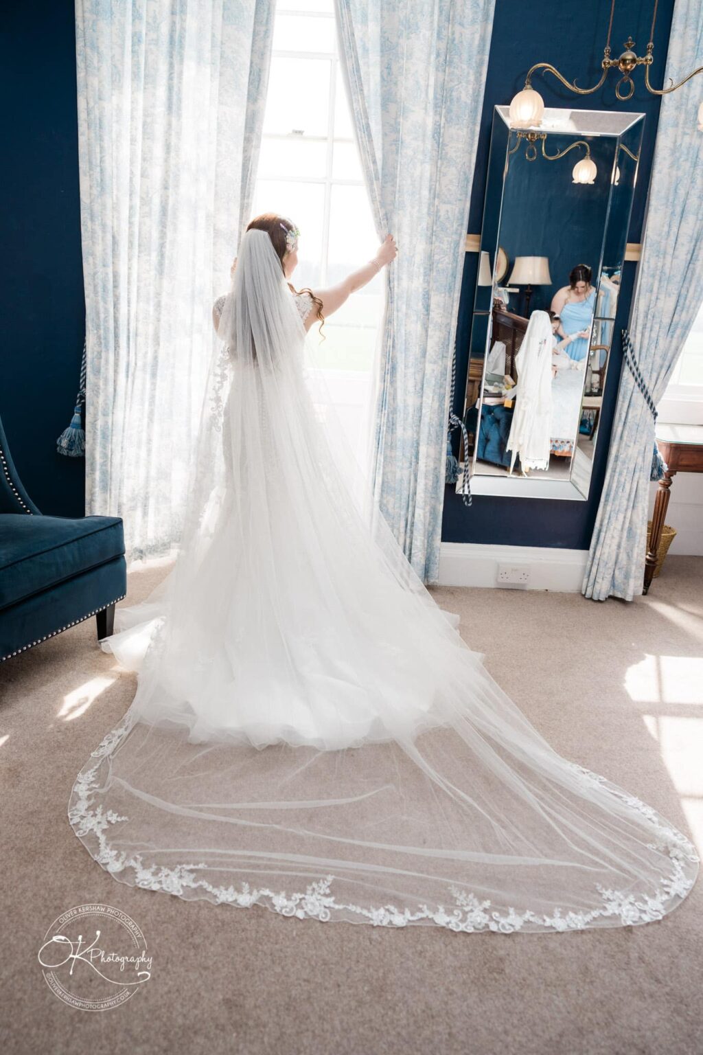 Prestwold Hall Wedding Photography Bride in a flowing white gown and veil standing by a window, holding open curtains. A large mirror on the side reflects her image. The room has a blue interior with elegant decor.