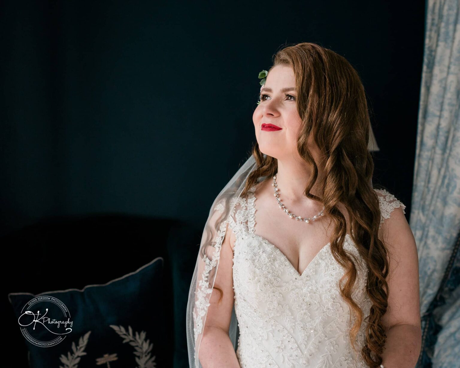 Prestwold Hall Wedding Photography A bride with long wavy hair wearing a lace wedding dress and pearl necklace, standing with a thoughtful expression indoors.