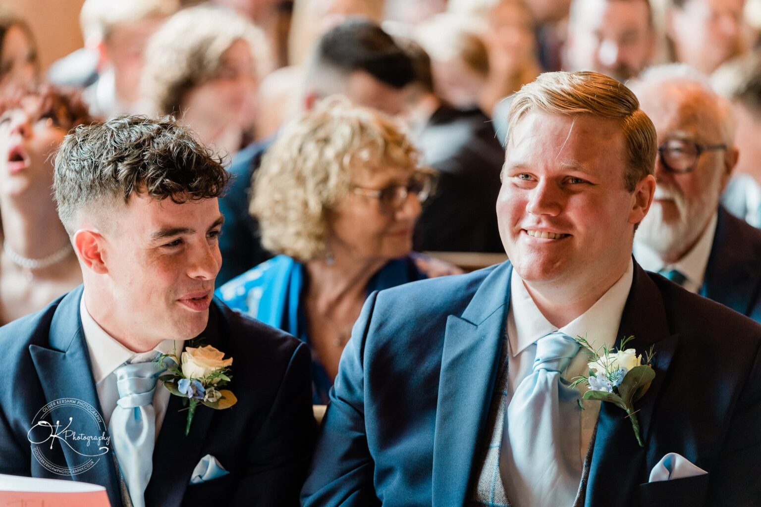 Prestwold Hall Wedding Photography Two men in suits, adorned with boutonnières, smiling and talking among a crowd at what appears to be a formal event or wedding.