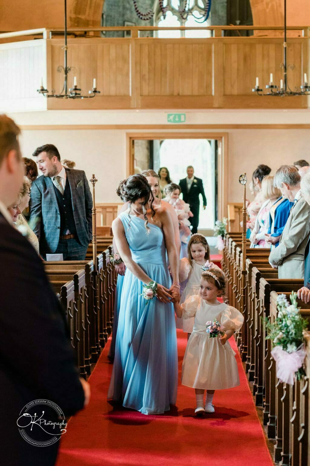 Prestwold Hall Wedding Photography Bridesmaids and flower girls walking down the aisle at a church ceremony, with guests seated on either side.