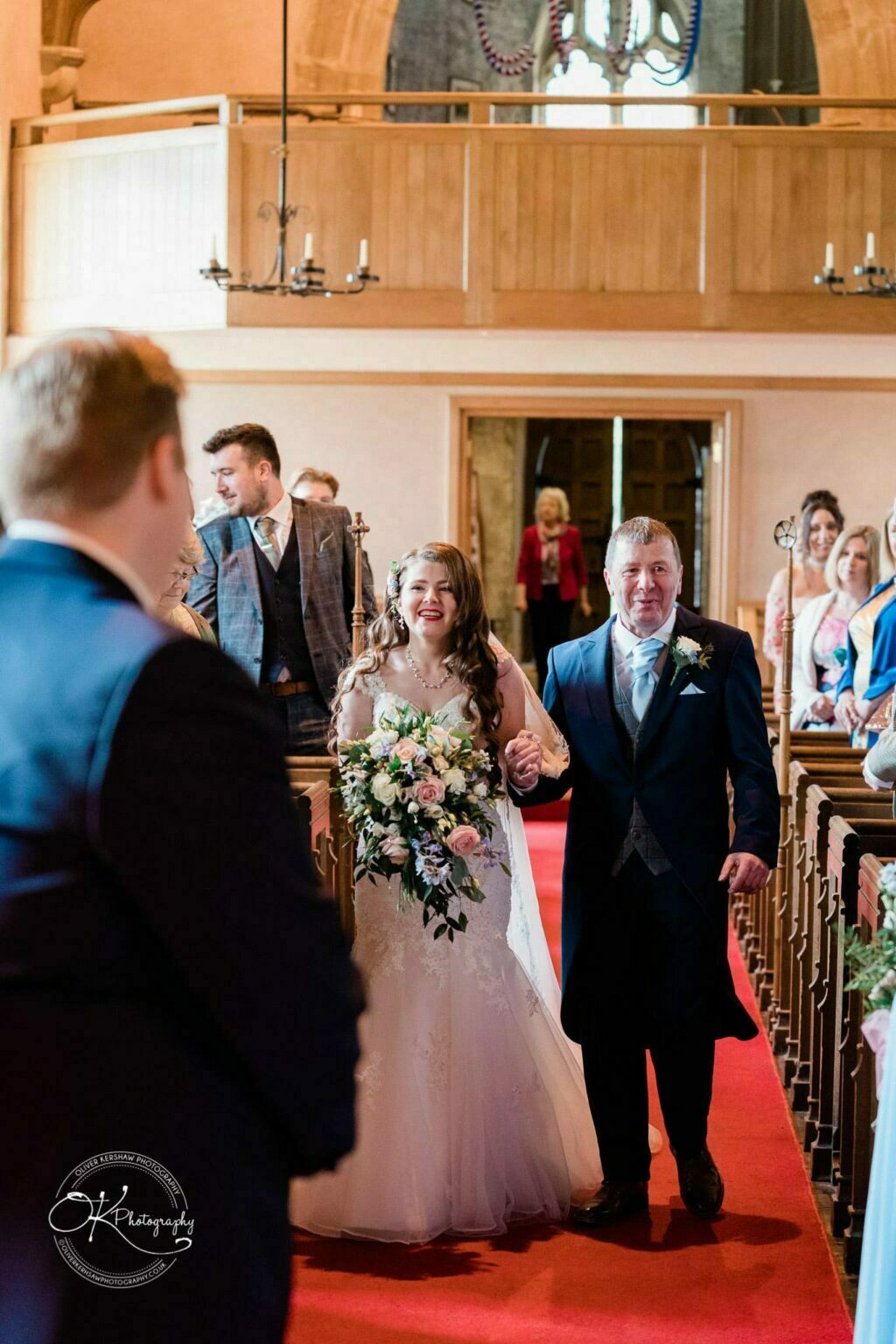 Prestwold Hall Wedding Photography A bride, escorted down the aisle by her father, holds a bouquet of flowers while guests look on in an elegant, wood-paneled hall.