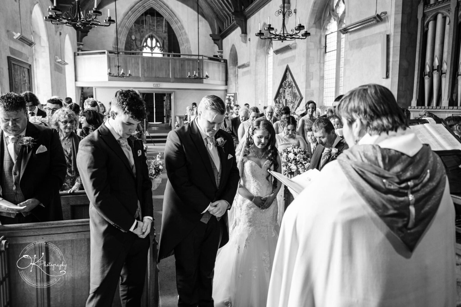 Prestwold Hall Wedding Photography A wedding ceremony inside a church, with the bride and groom standing at the altar, along with groomsmen and guests seated in the pews.